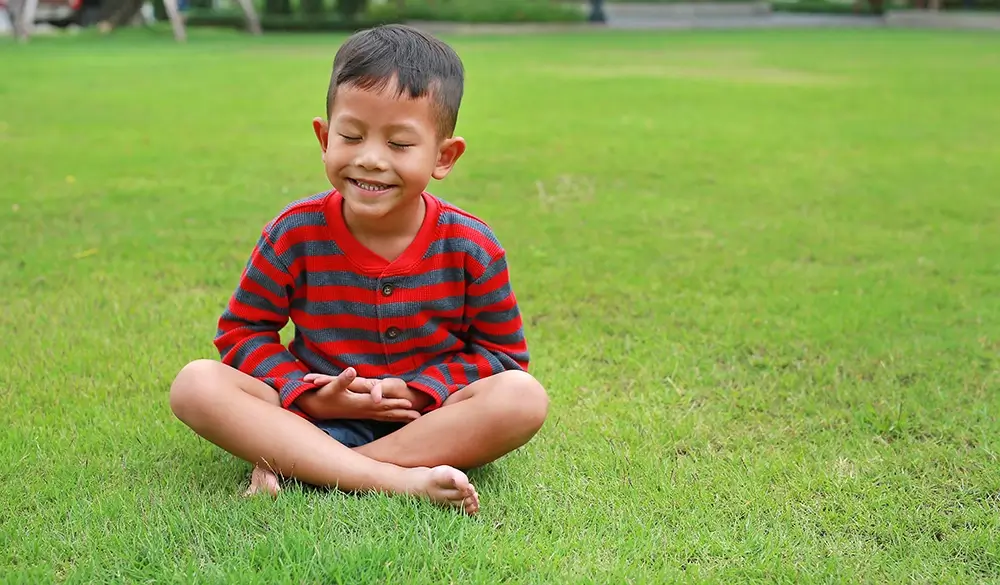 Young asian boy sits in the grass meditating and smiling.